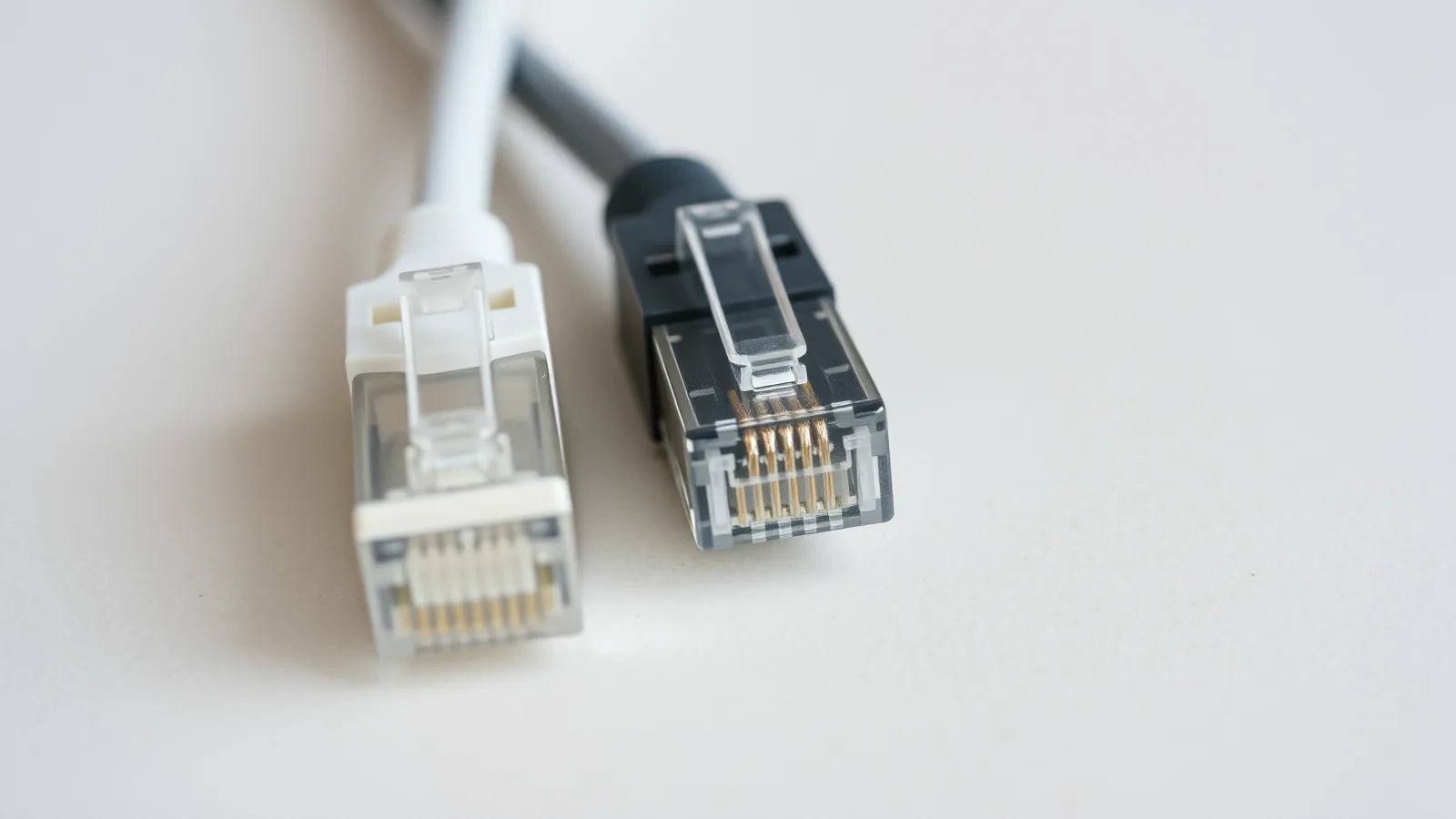 Close-up van een witte en een zwarte netwerkkabel met doorzichtige RJ45-stekkers op een licht werkblad